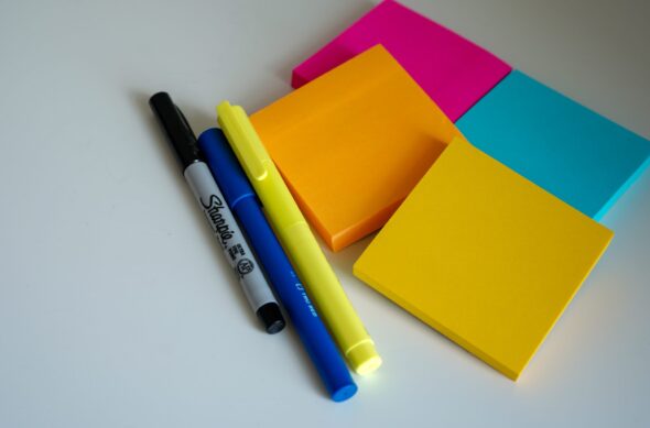 A pen, marker, and highlighter next to four stacks of sticky notes in orange, yellow, bright pink, and blue.